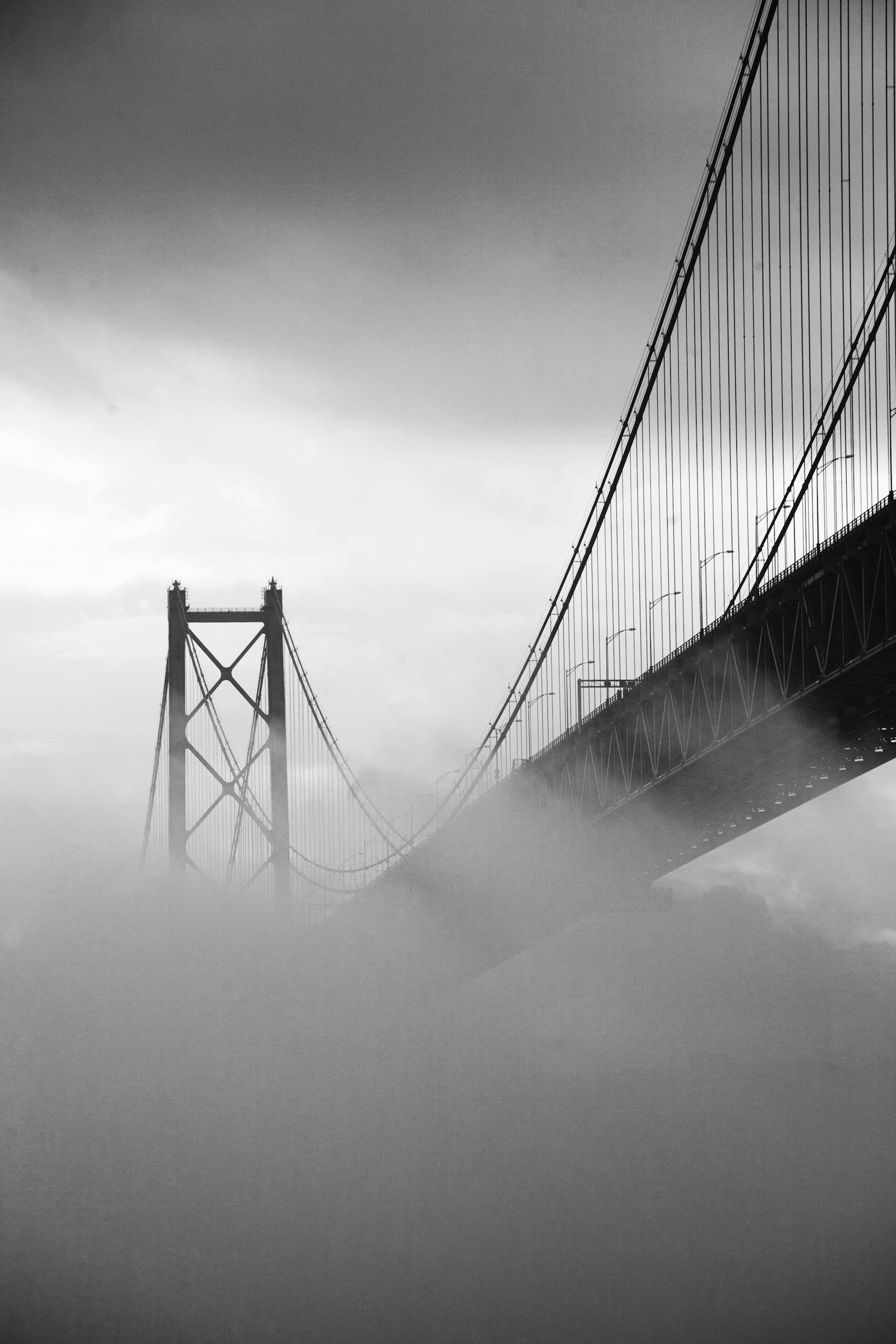 Black and white photo of a suspension bridge enveloped in fog, creating a dramatic scene.